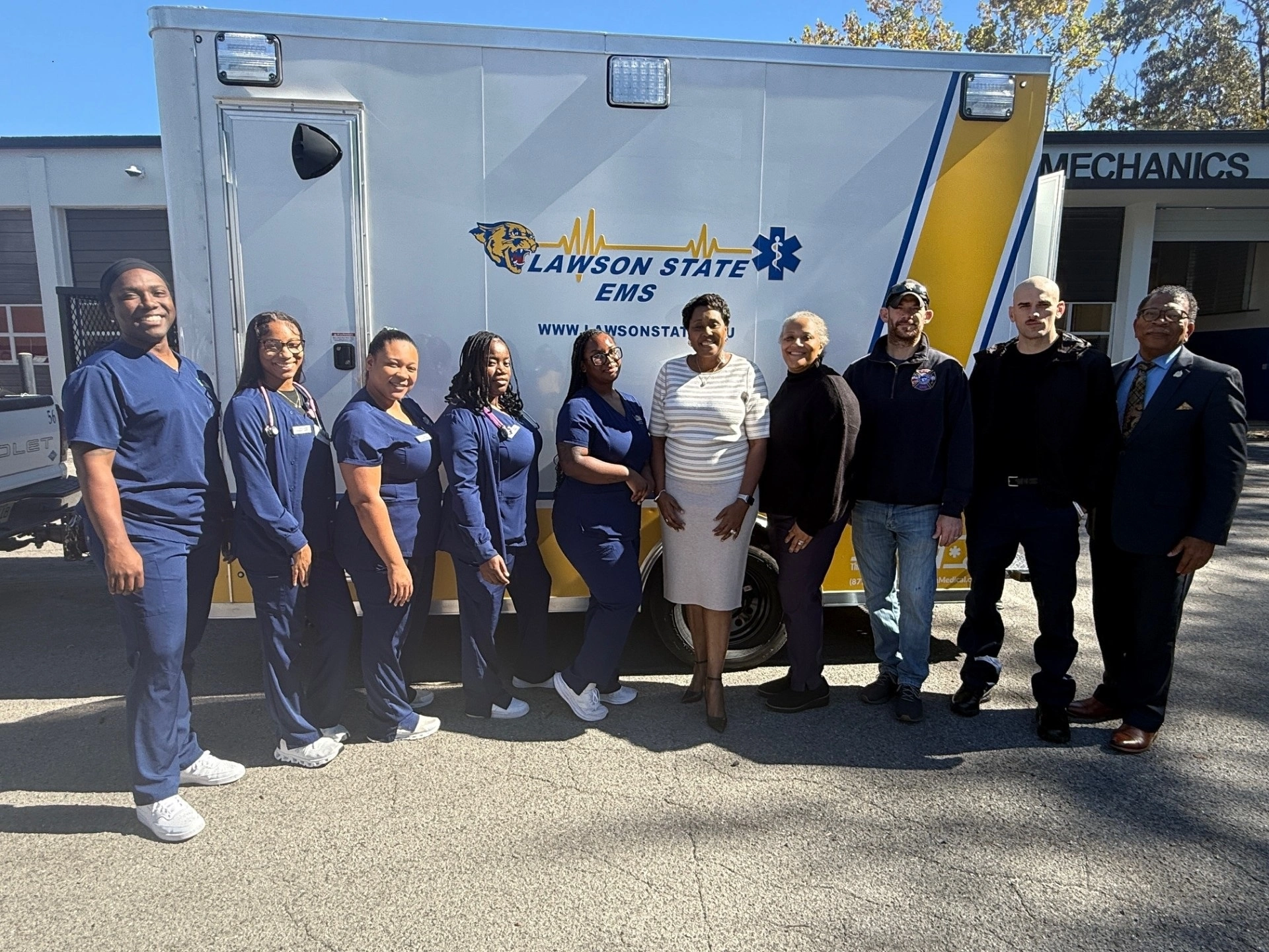 Students from various health professions programs pose with President Cynthia T. Anthony in front of the college’s new yellow, blue and white SimRig, a mobile emergency medical services trailer that will enhance student learning.