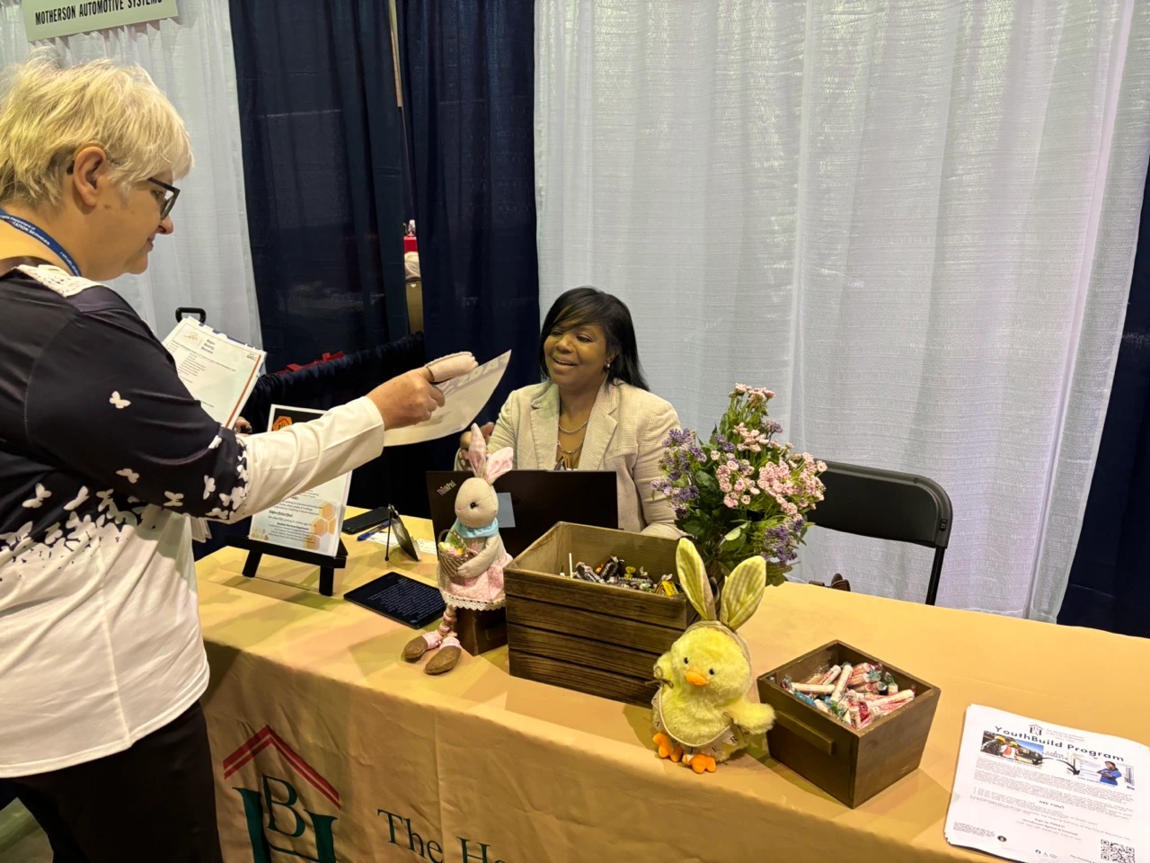 A booth at a community fair featuring a table covered with spring‑themed decorations, stuffed animal figures, wooden boxes filled with candy, informational flyers, and a small bouquet of flowers. One woman stands at the table engaging with the booth representative seated behind it.