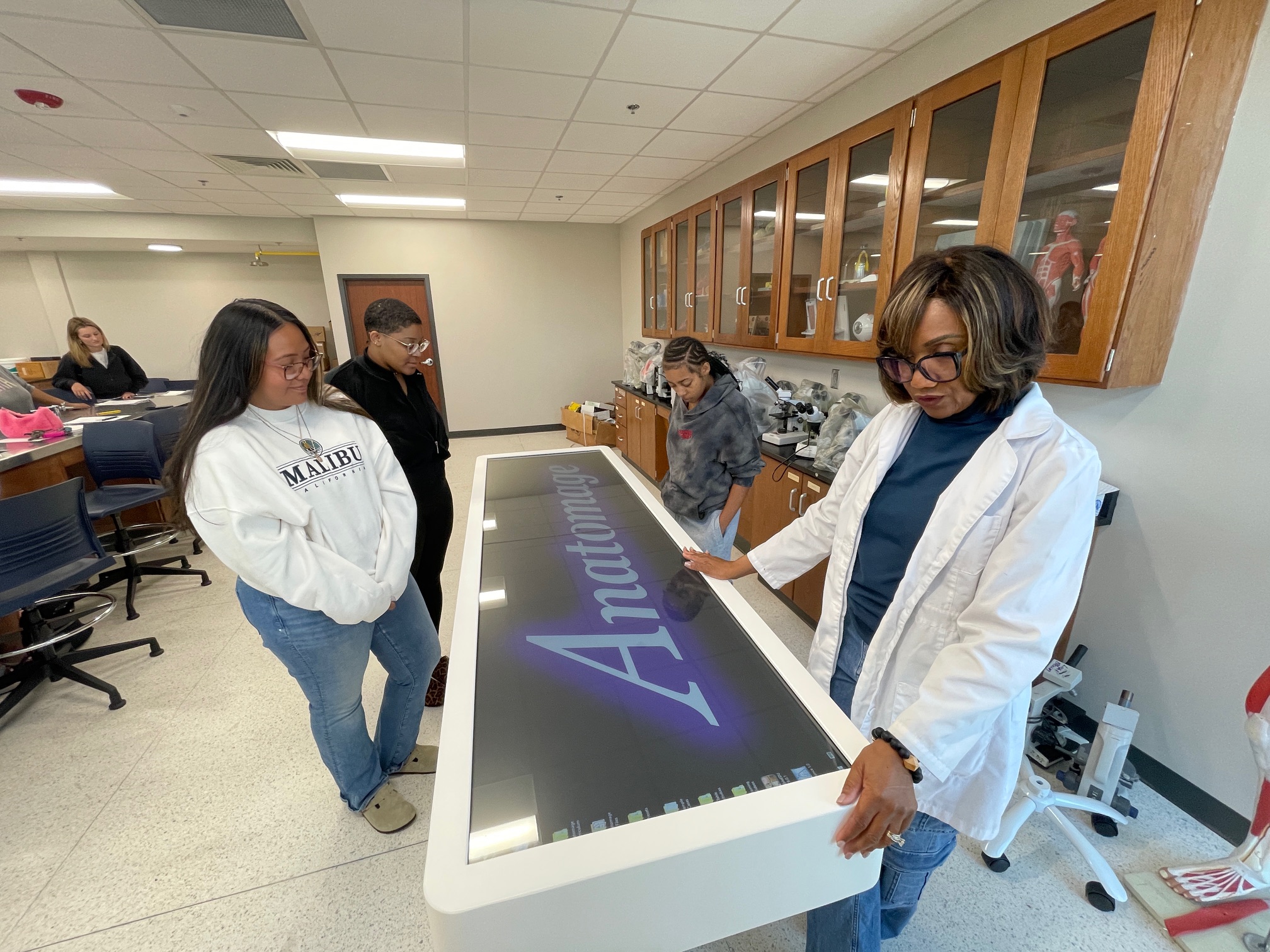 An instructor in a white lab coat demonstrates the Anatomage virtual dissection table to students in a Lawson State science lab.