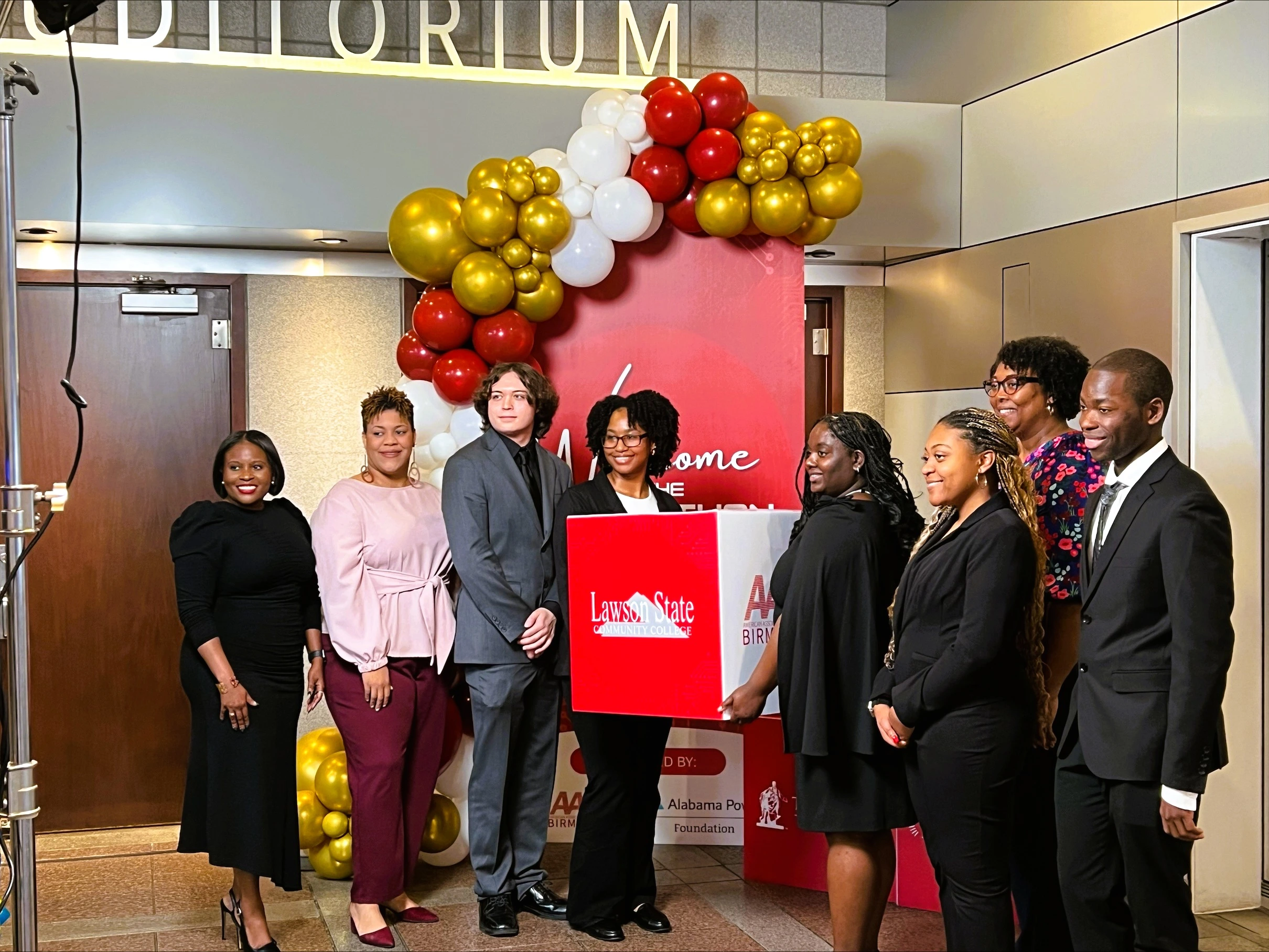 A group of eight professionally dressed people pose and smile in front of a red Lawson State Community College backdrop with a red, white, and gold balloon arch.