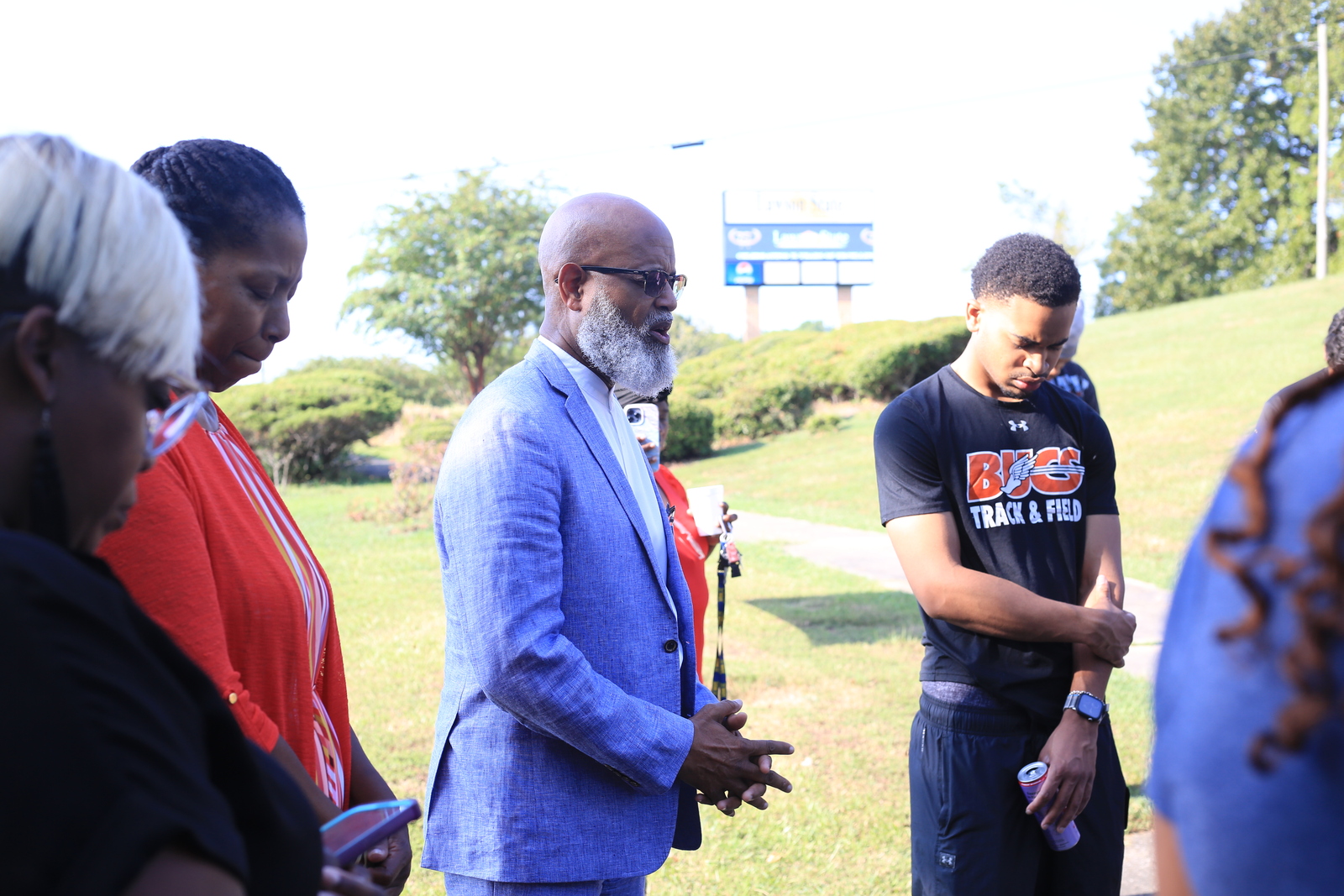 Lawson State students, faculty, and staff bow their heads in prayer on the campus lawn as a speaker in a blue suit leads the group, with a student in a Bucs Track & Field shirt listening nearby.