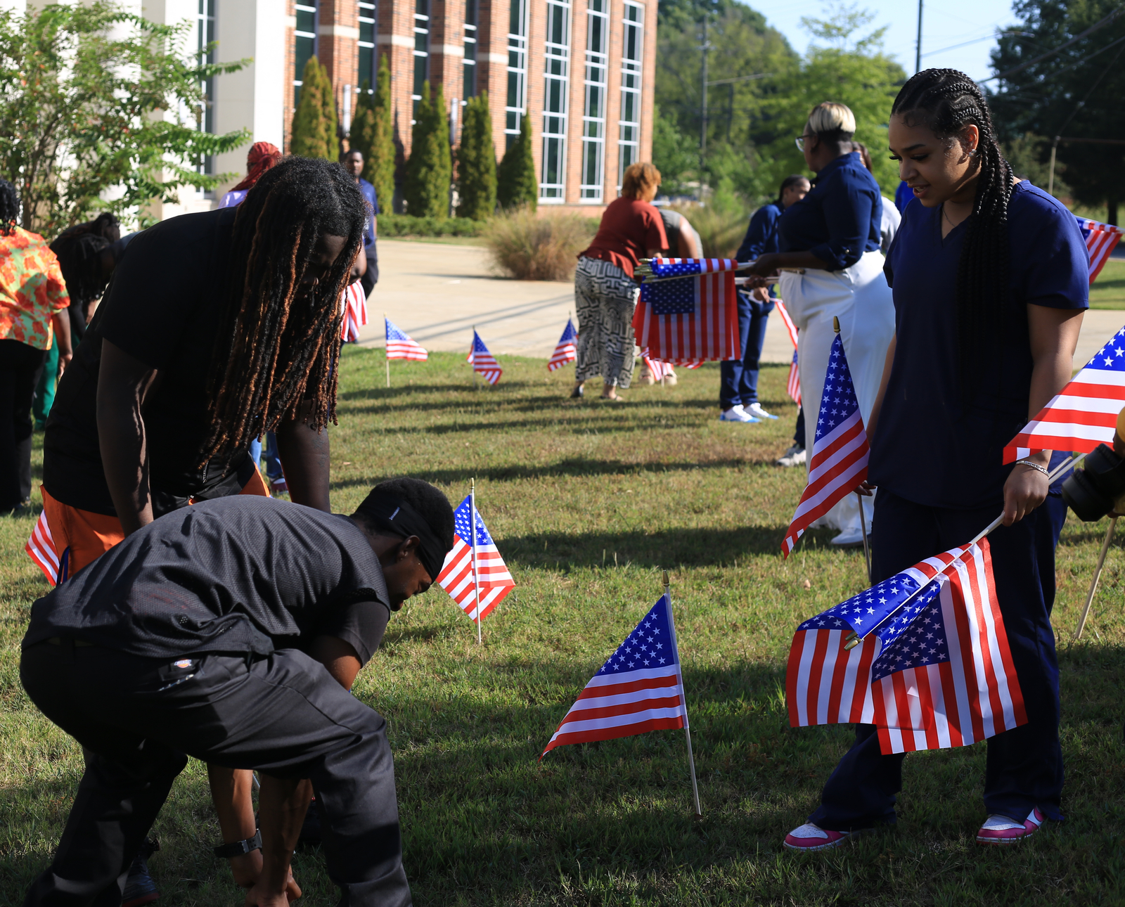 Lawson State students and staff place small American flags in the lawn in front of a brick campus building, with one student carrying an armful of flags and others kneeling to plant them.
