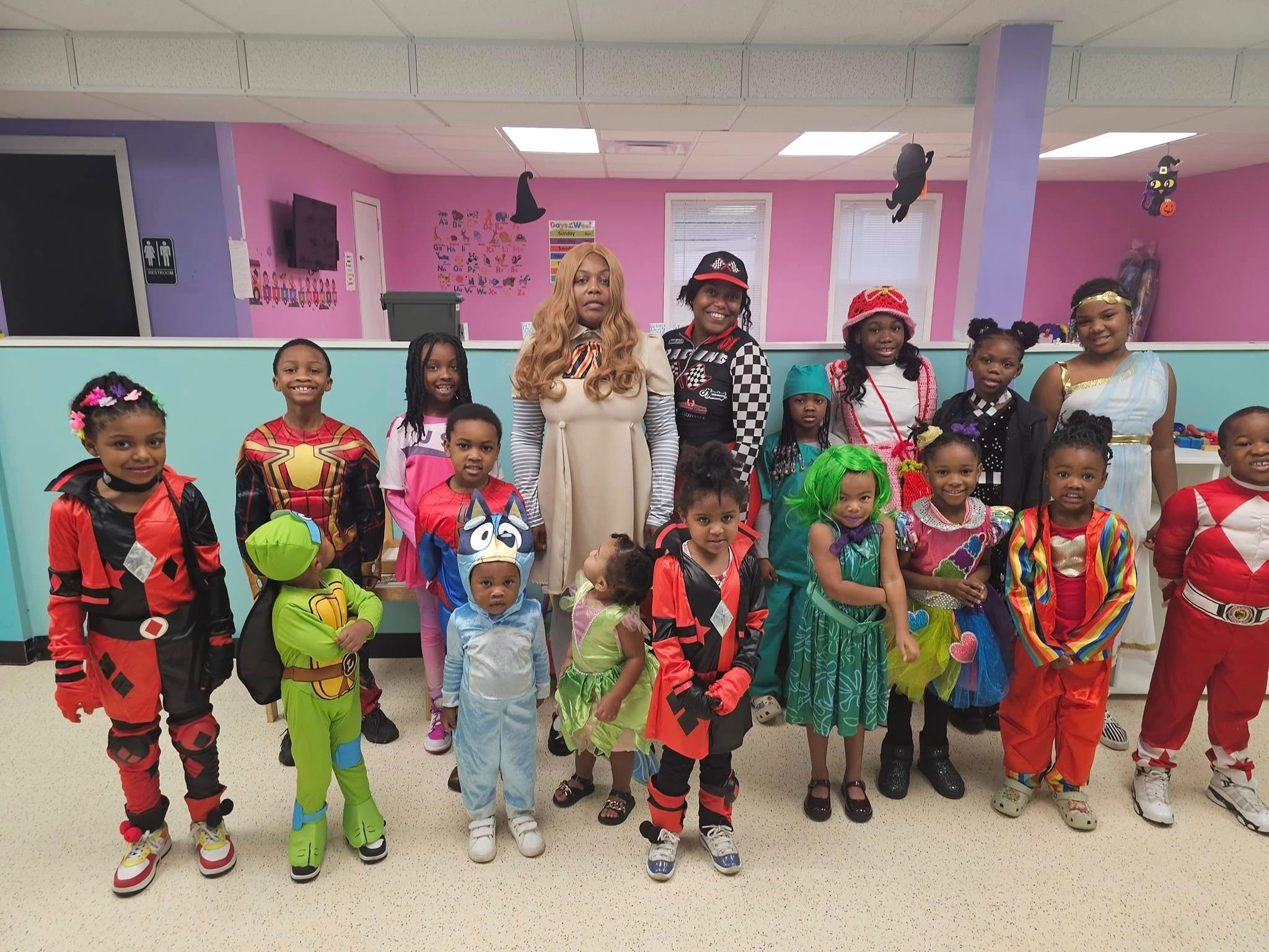 Preschool children dress in Halloween costumes like ninja turtles, power rangers, clowns, and other characters facing the camera with daycare teachers smiling behind them. 