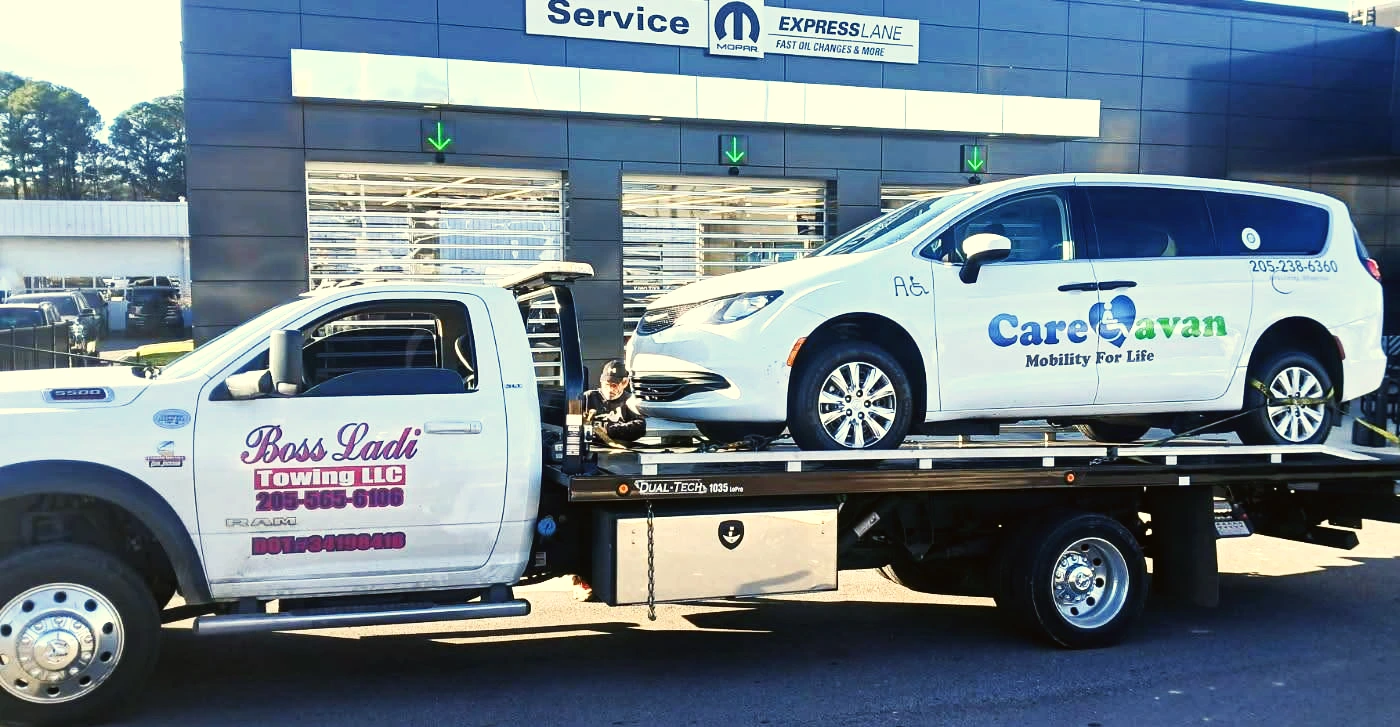A white flatbed tow truck with pink logo hauls a nonworking white minivan to a automotive service center for repairs. 