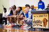 Coach crouches courtside near a 'Home of the Lawson State Cougars' sign, focused on the game while players sit blurred on the bench behind