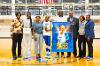 A Lawson State volleyball senior wearing jersey number 12 stands on the gym court holding flowers, surrounded by family, coaches, and staff, with a large poster reading "Zoe Rogers" displayed in front.