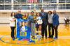 A Lawson State volleyball senior stands on the court holding flowers and a gift basket, surrounded by family, coaches, and staff, with a large poster reading "Jasmine Tyler" displayed in front.