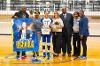 A Lawson State volleyball senior wearing jersey number 16 stands on the court holding a gift basket and flowers, surrounded by family and staff, next to a large poster reading "Mackenzie Richards."