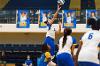 A Lawson State volleyball player jumps to set the ball near the net during a match, while teammates in blue-and-white uniforms prepare around her on the court.